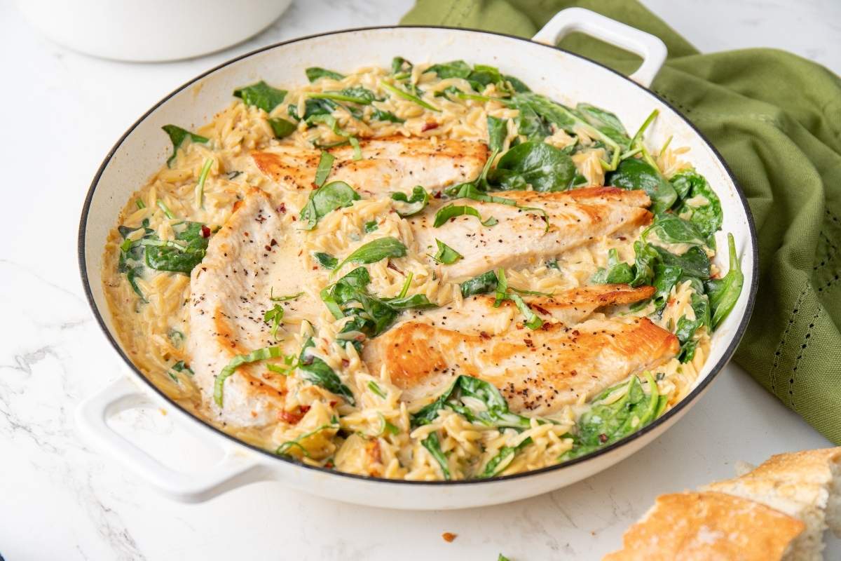 A white skillet containing creamy Tuscan Italian Marry me chicken orzo with cooked chicken breast slices atop creamy pasta and spinach, positioned on a marble surface next to a green cloth and a slice of bread.