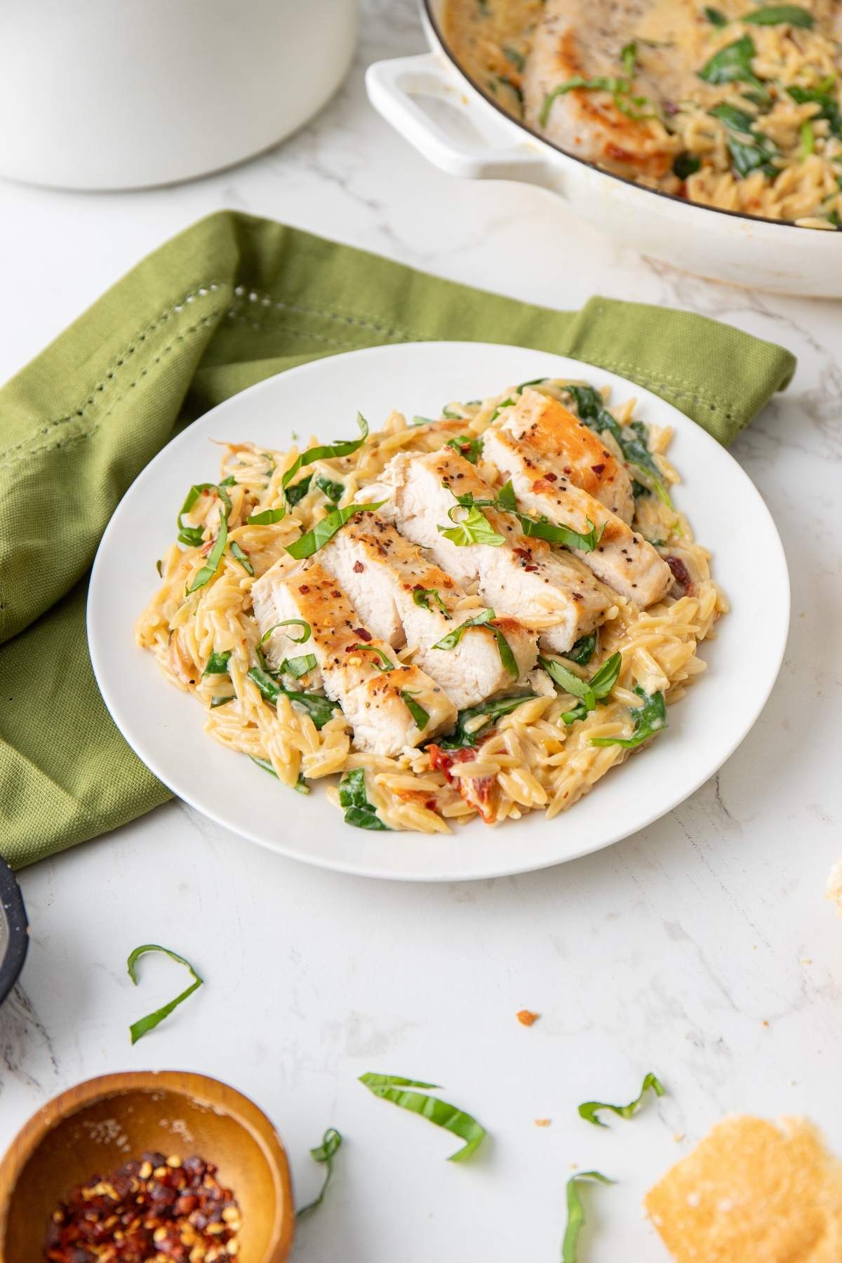 A white plate with Italian chicken orzo featuring grilled chicken slices on a bed of orzo pasta mixed with spinach, garnished with herbs, placed on a green napkin. A pot and additional ingredients are visible in the background.