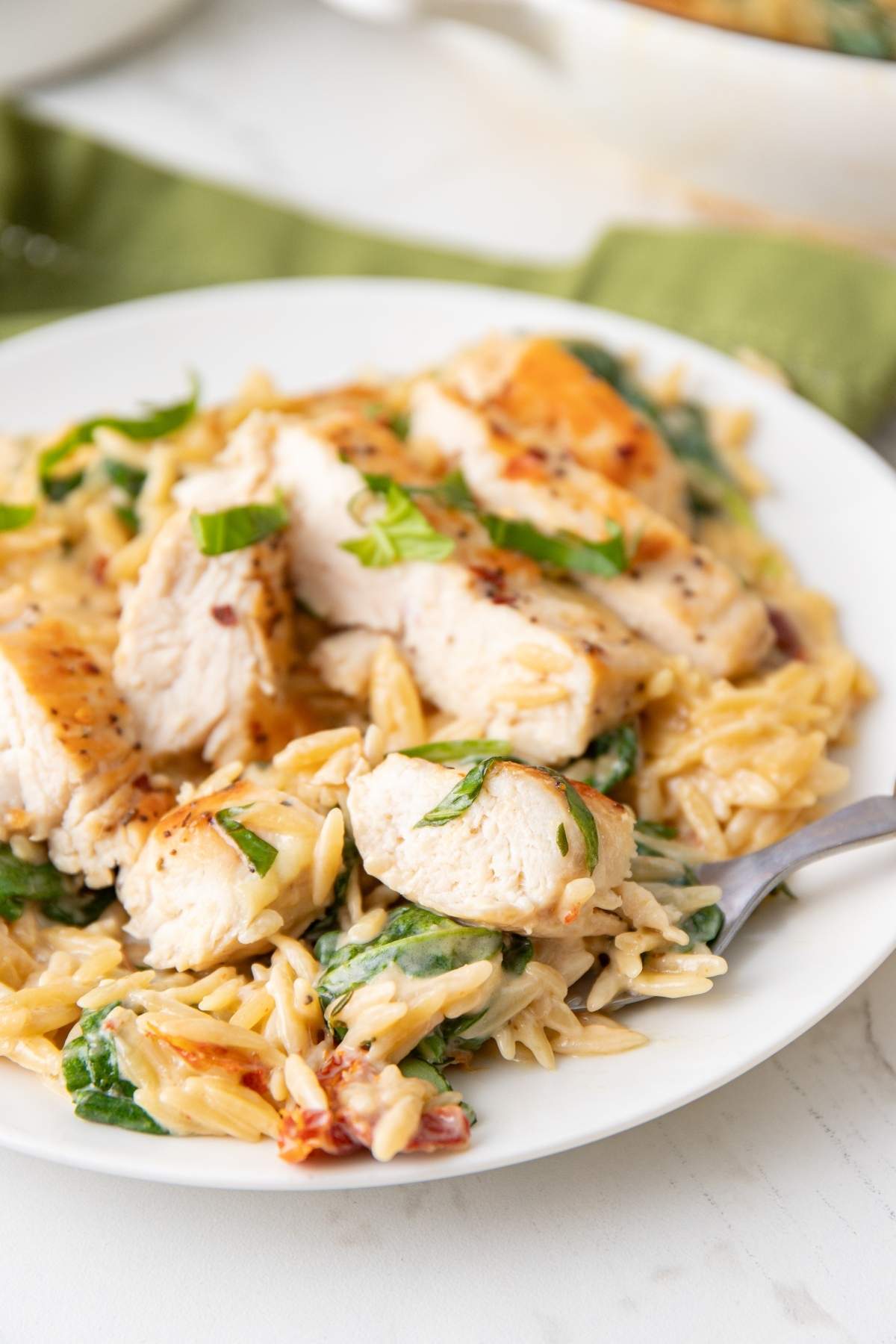 A white plate with Italian chicken and orzo, featuring sliced chicken breast served over creamy orzo pasta mixed with spinach and garnished with herbs, with a fork resting on the side.