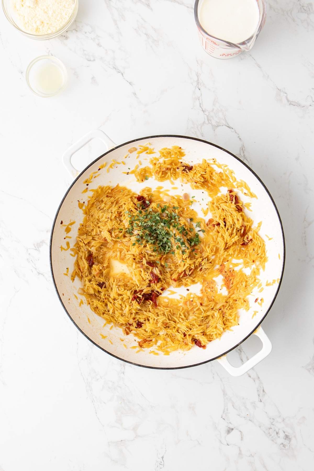 A white pan containing sautéed rice and chopped herbs, likely parsley, atop a white marble surface, with bowls of various ingredients including Italian chicken orzo and grated cheese in the background.